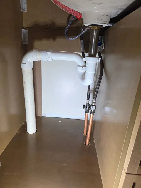 Underside view of a kitchen sink area showing plumbing pipes, drain connections, and flexible water supply lines, with a brown cabinet and a wall in the background.