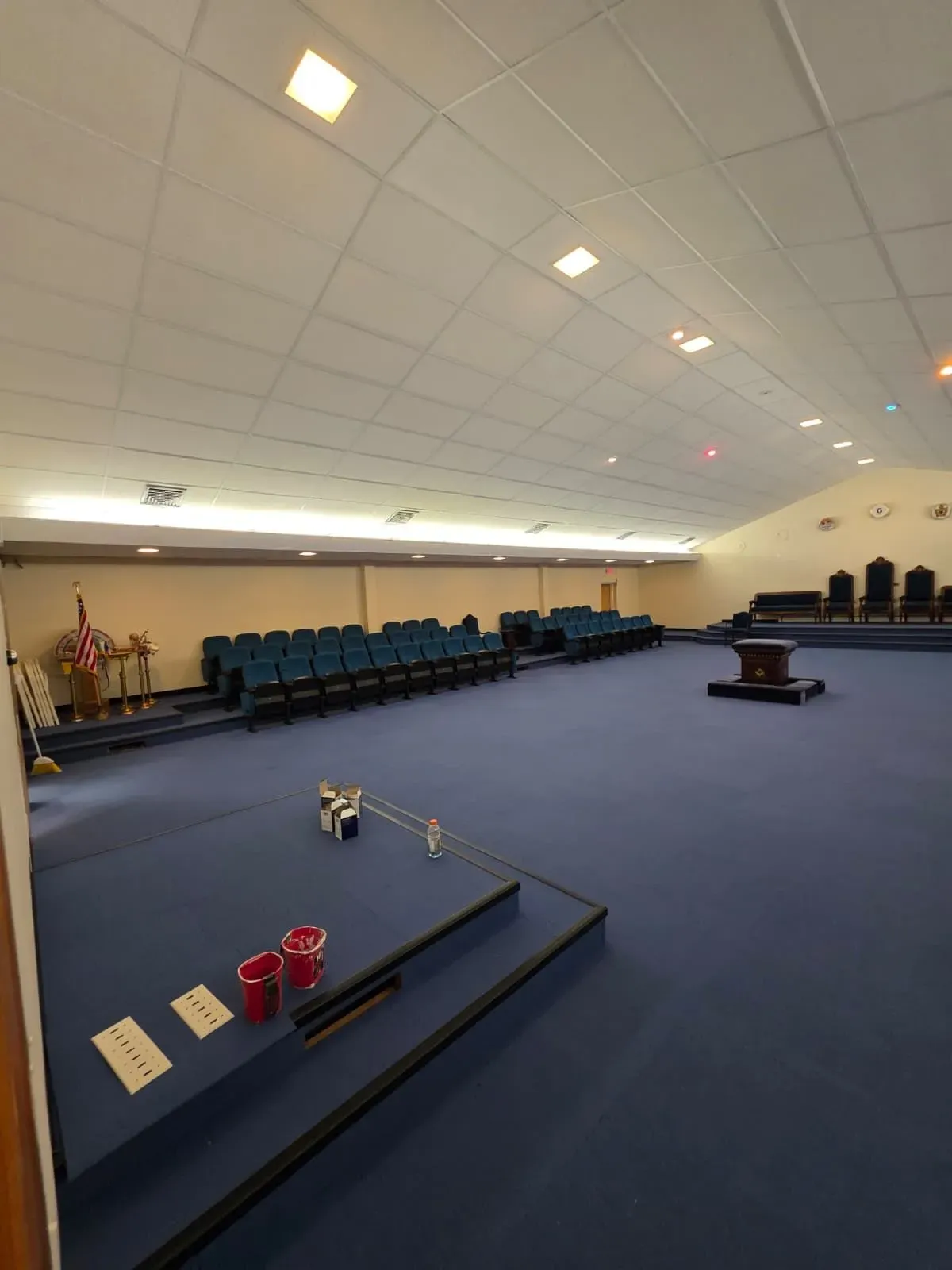 Empty church sanctuary with blue carpet, rows of teal chairs, flags, and a pulpit stage