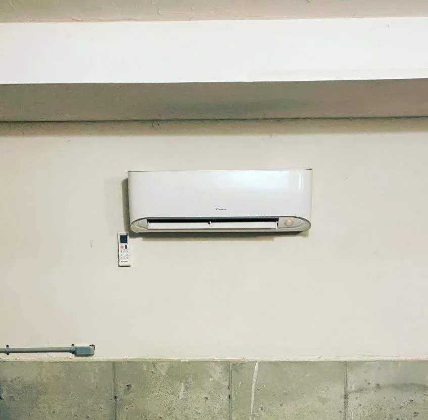 Wall-mounted white air conditioner with remote control on light-colored wall above concrete base.