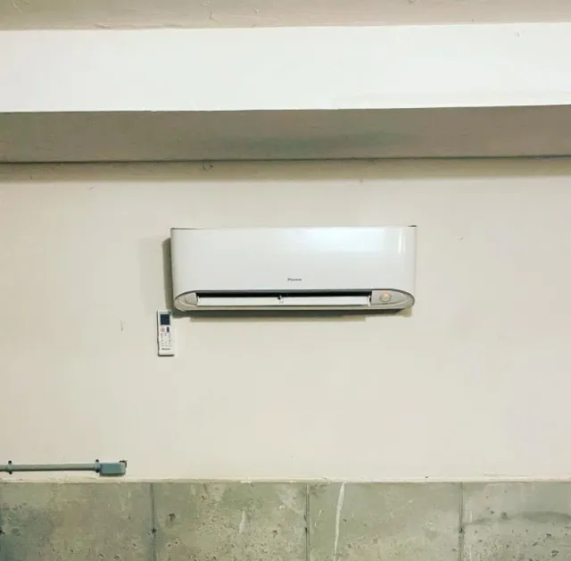 Wall-mounted white air conditioner with remote control on light-colored wall above concrete base.