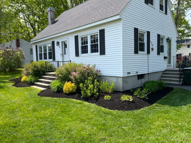 White house with black shutters, stone steps leading to the front door, green grass, and landscaped garden with bushes and flowers.