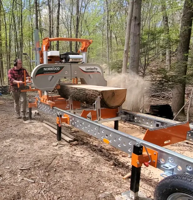A person operating a large portable chainsaw mill cutting through a fallen log in a wooded forest.