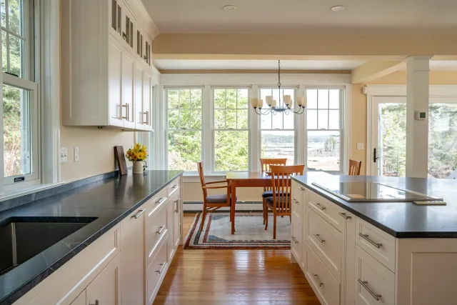 Bright kitchen with white cabinets and black countertops, adjoining dining area with wooden table and chairs, large windows overlooking trees, and a chandelier above the table.