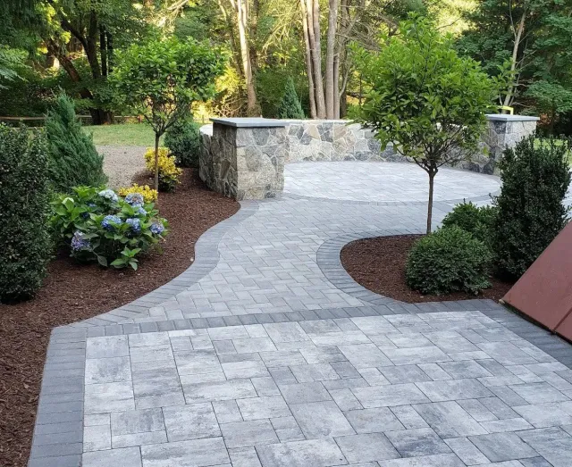 Curved gray stone patio path through a landscaped garden with shrubs, trees, and a stone wall