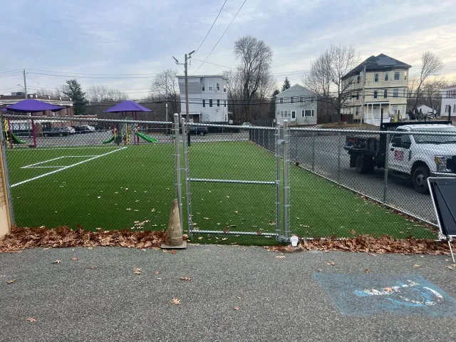 Fenced-in area with artificial turf, gate open, residential buildings in background, cloudy sky.