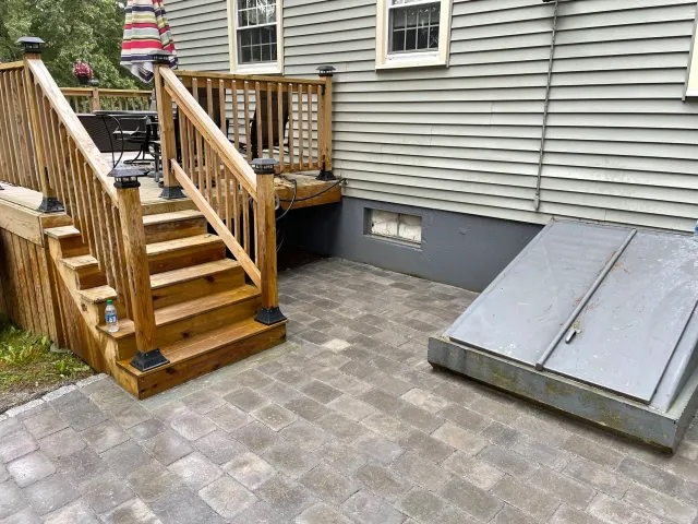 Outdoor wooden deck and stairs with a table and chairs; adjacent to a house with grey siding and a metal basement door on a brick patio.