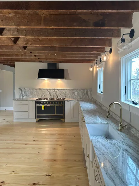 Kitchen with wooden ceiling beams, white cabinets, a marble countertop, a black stove and range hood, and a brass faucet over a white sink. There are three windows with a view outside, and a row of modern light fixtures along the wall.