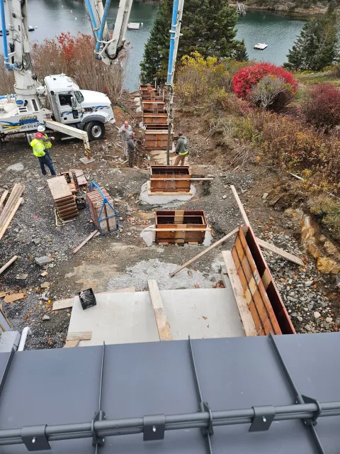 Construction workers and a concrete truck are building a pathway near a body of water, with colorful trees and shrubs in the background.