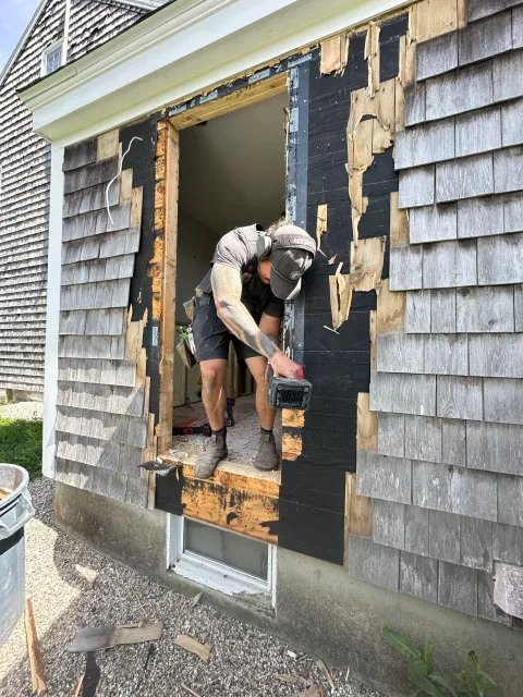 A man working on a house removal or renovation, using a power tool at a large open window frame. The house exterior has weathered wooden shingles, and construction debris is visible on the ground.