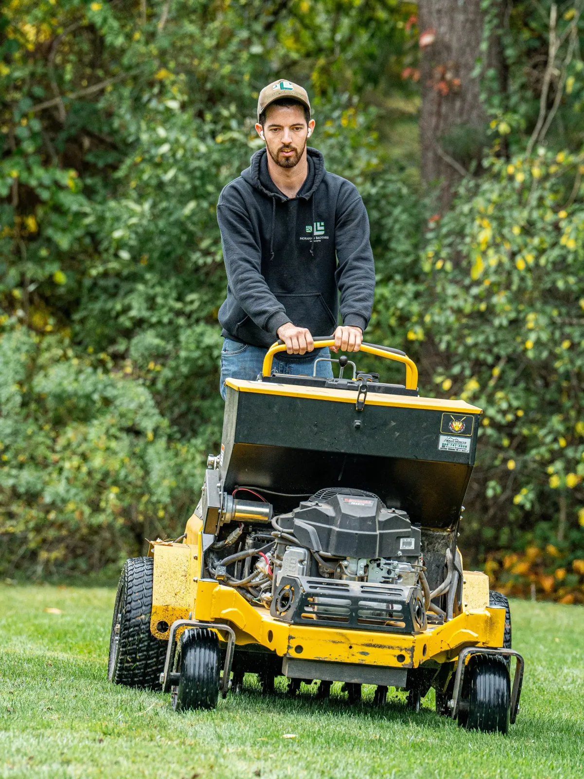 A man wearing a black hoodie and a beige cap is operating a yellow turf aerator on a grassy lawn, with bushes and trees in the background.