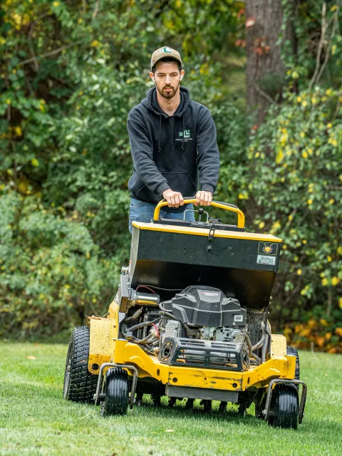 A man wearing a black hoodie and a beige cap is operating a yellow turf aerator on a grassy lawn, with bushes and trees in the background.