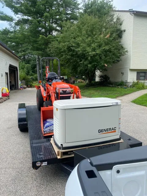 Orange compact tractor towing a white cooler on a gravel driveway near houses and trees