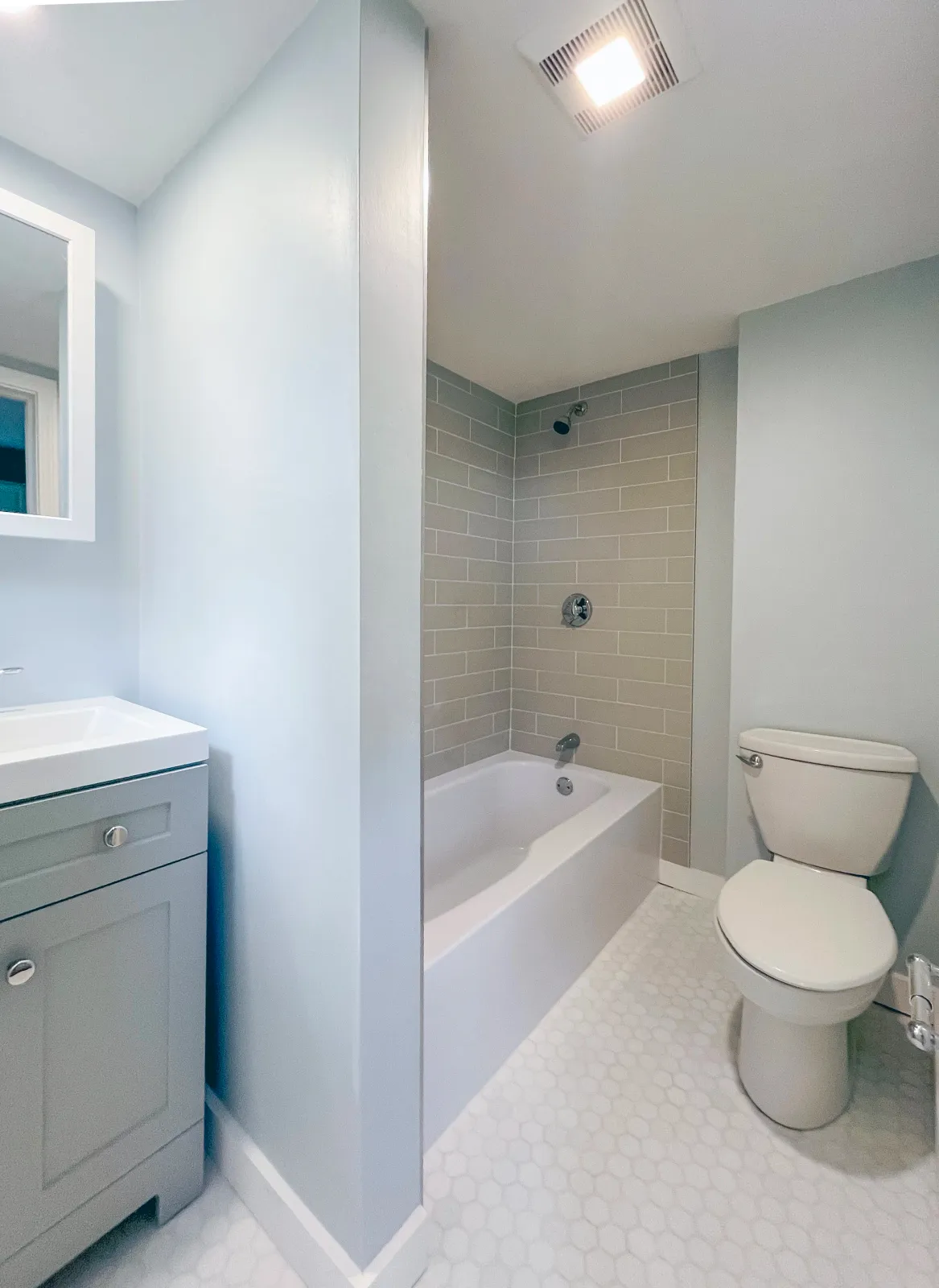 A small bathroom with a vanity, mirror, bathtub with beige tiles, and a toilet. The walls are painted light blue, and the floor has hexagon white tiles.