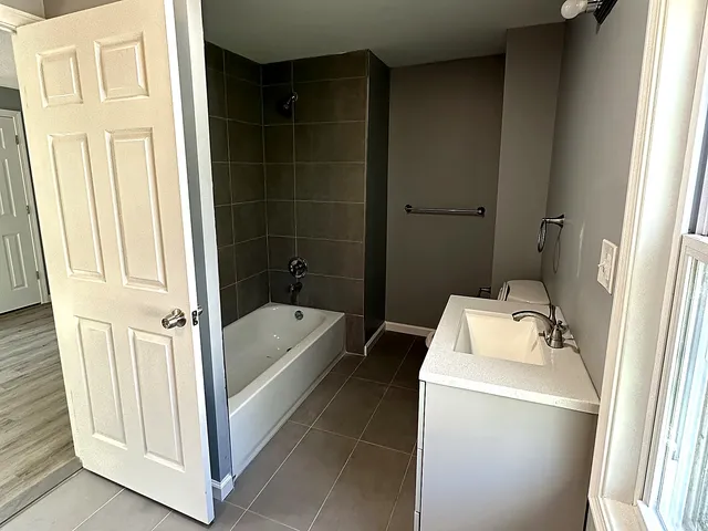 Remodeled bathroom in Watertown Connecticut with large-format grey porcelain floor tile, a charcoal tile tub and shower surround with chrome trim, a white soaking tub, a white vanity with a solid-surface top and chrome faucet, warm grey walls, chrome vanity lighting, and a new ceiling exhaust fan and light