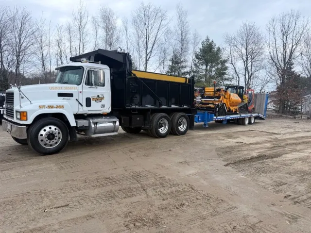 A flatbed truck parked outdoors on a dirt surface, carrying construction equipment including bulldozers and excavators, with leafless trees in the background.