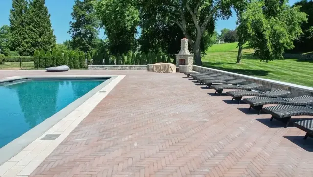 Empty swimming pool with a brick patio, lounge chairs, a stone outdoor fireplace, and green trees in the background on a sunny day.