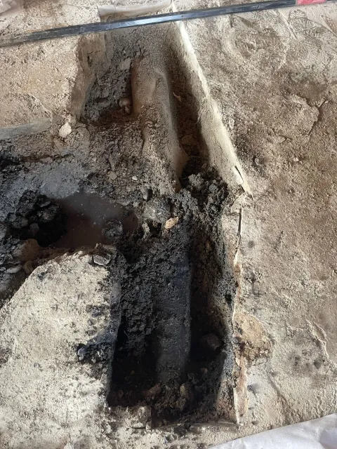 An excavation pit revealing dark soil and objects in a sandy environment, potentially an archeological site.
