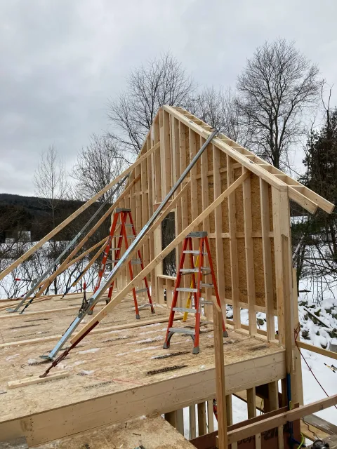 Wood framing of a house under construction with ladders and metal braces, snowy landscape with leafless trees in background.