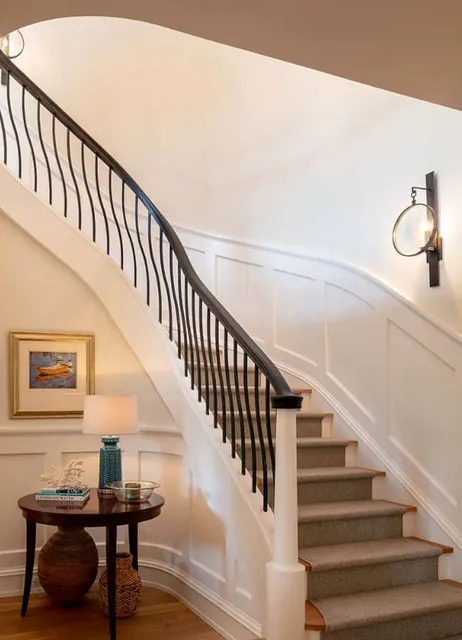 Elegant curved staircase with a gray stair runner installation, black wrought iron railing, and white wainscoting. A round side table with a blue lamp and artwork adds a touch of sophistication.