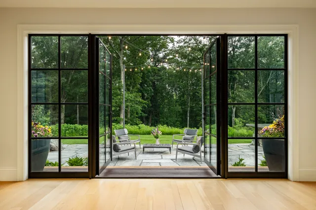 A spacious, well-lit room opens to a patio with furniture, string lights, and a lush green background through large black-framed glass doors.