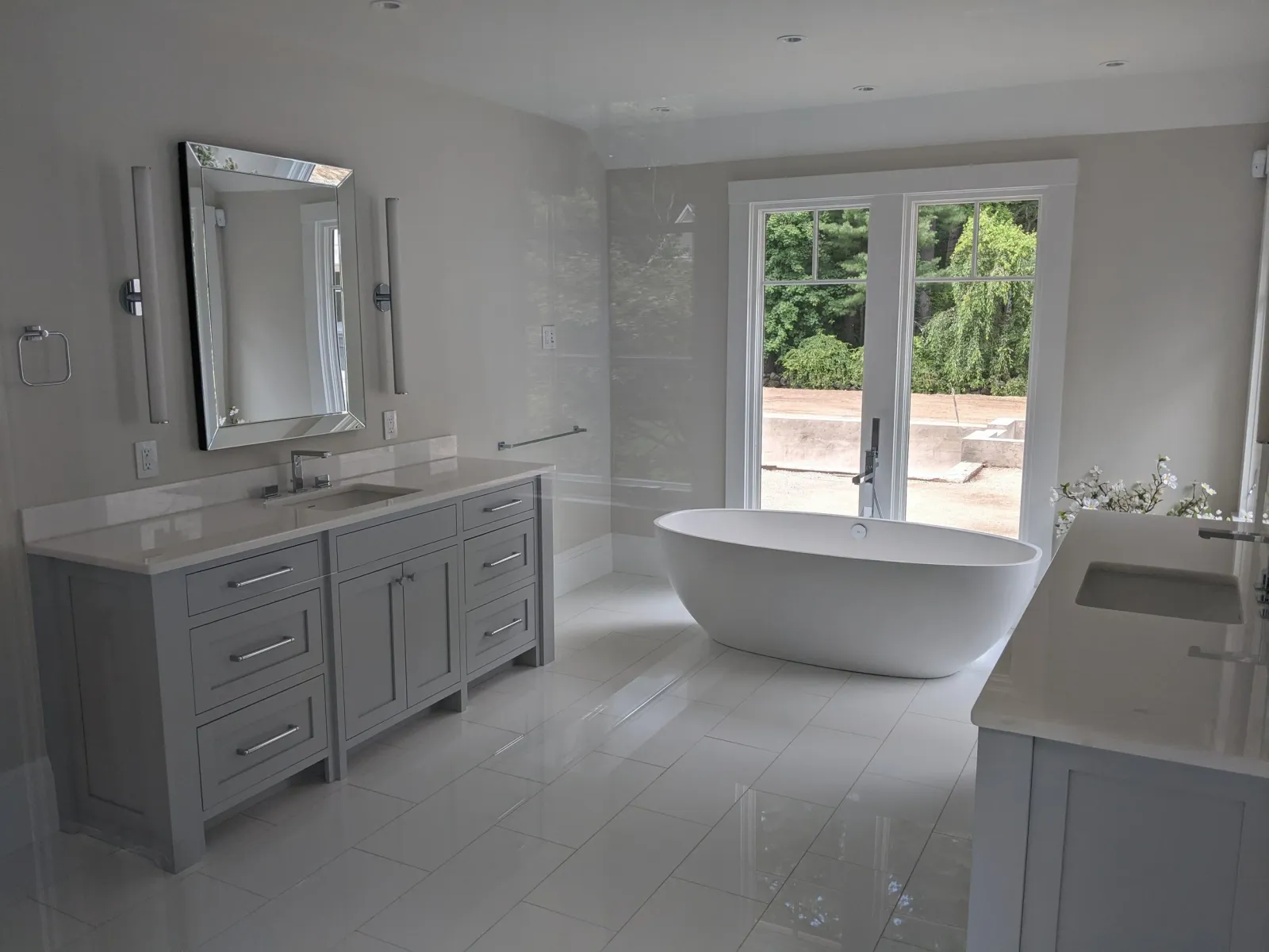 Modern bathroom with a white freestanding bathtub, double sink vanity, and a large window with a view of trees outside.