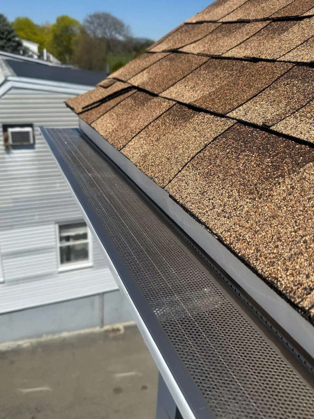 Detailed view of brown asphalt roof shingles and a newly installed metal gutter guard with a fine mesh screen, designed to prevent debris buildup, on a clear sunny day above a residential neighborhood.