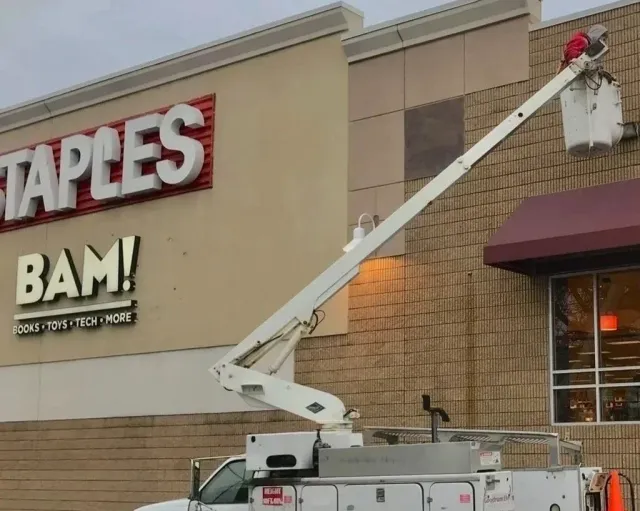 A white utility bucket lift truck is positioned outside a Staples store, with the basket raised to the store's exterior wall.