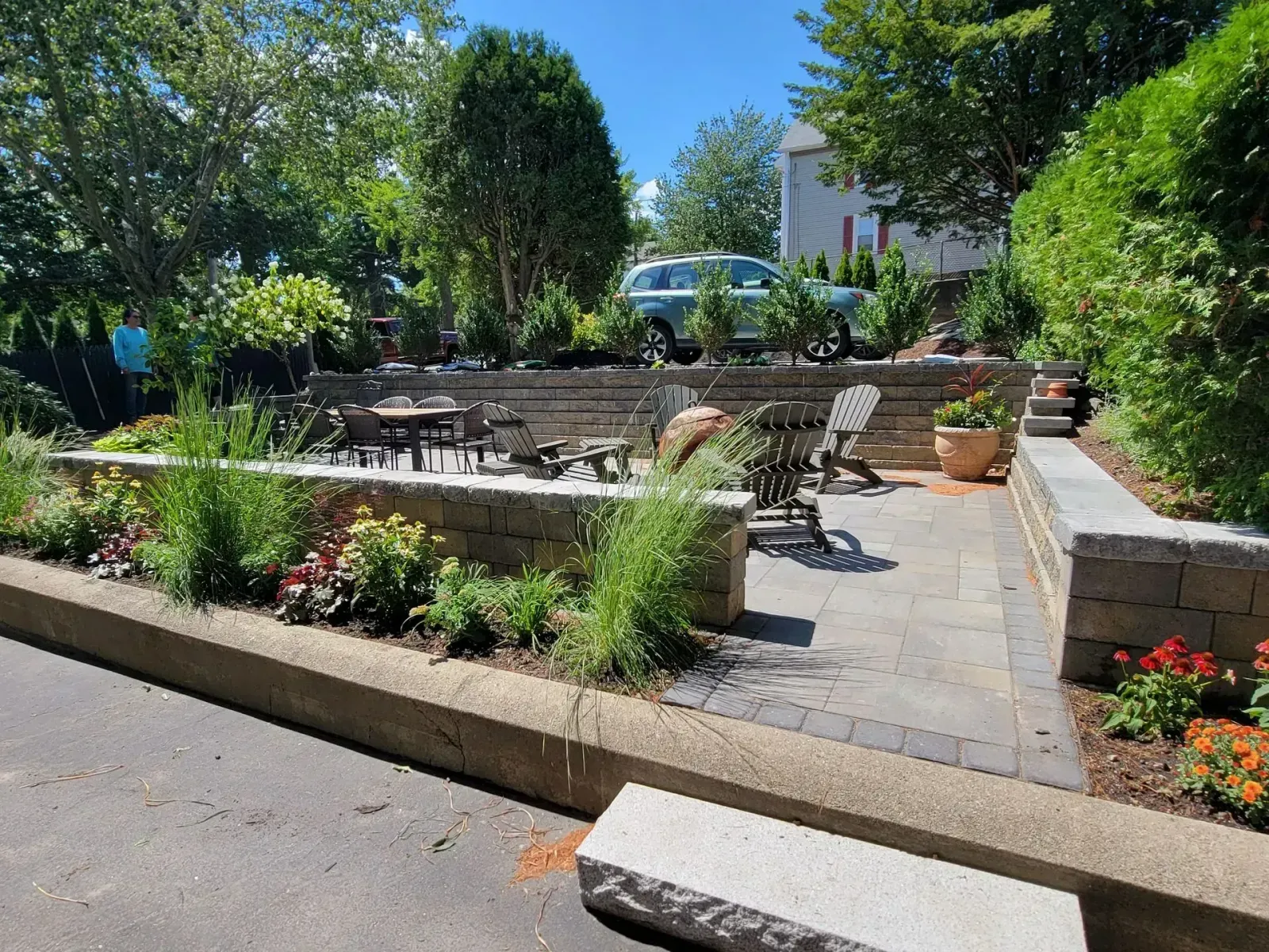 A fire pit with white Adirondack chairs on a stone patio in a yard, surrounded by mulch and lush trees.