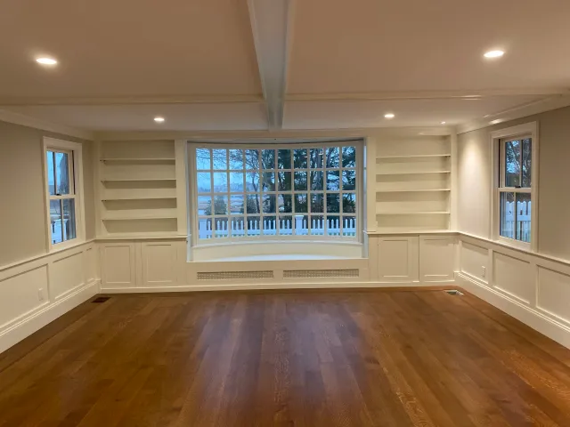 Empty living room with large bay window, built-in bookshelves on both sides, hardwood flooring, and recessed ceiling lights.