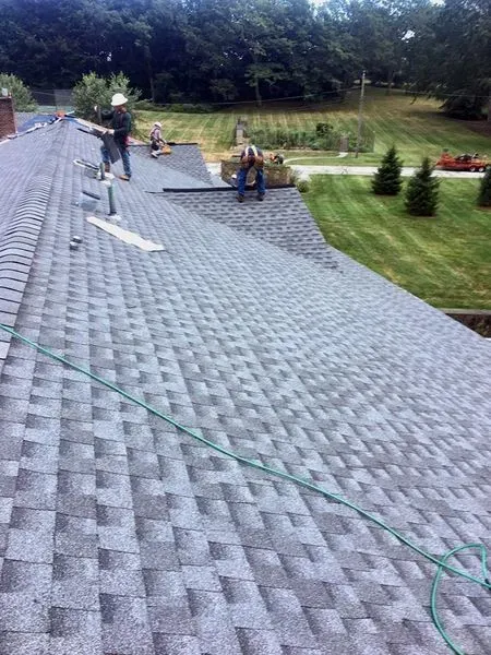 Roofers working on a gray shingled roof near a chimney and trees in a residential setting.
