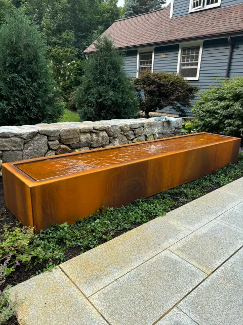 A Corten steel fountain is a focal point and adds the cooling sound of water to this granite patio project. Adjacent landscape design includes a field stone knee wall, a fire bowl area, vegetable gardens and border plantings.