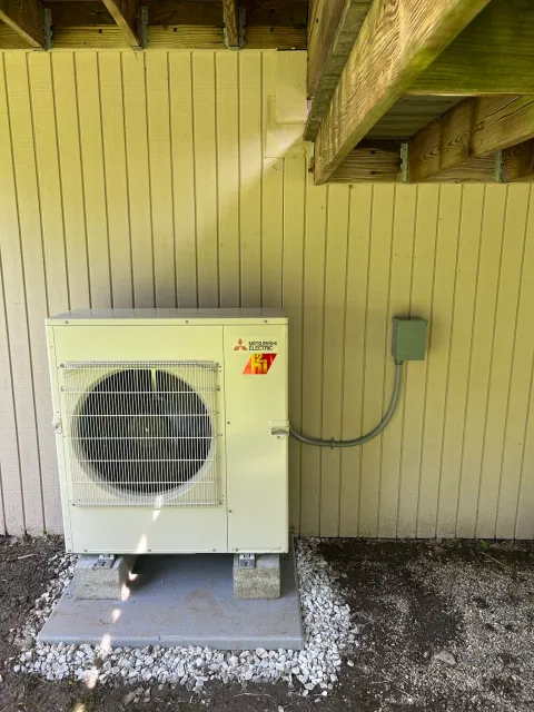 Outdoor Mitsubishi Electric HVAC unit on a concrete pad with gravel, next to a yellow wall and wooden porch above.