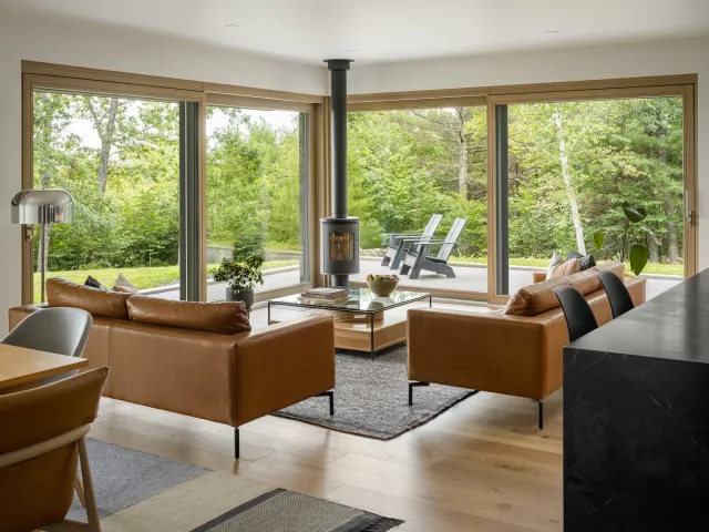 Living room with large windows overlooking a wooded outdoor space, tan leather sofas, a glass coffee table, a black wood stove, and indoor plants.