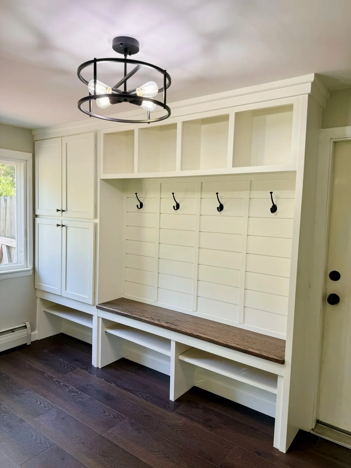 Kitchen mudroom with white built-in storage, black hooks, a wooden bench, a window, and a modern chandelier.