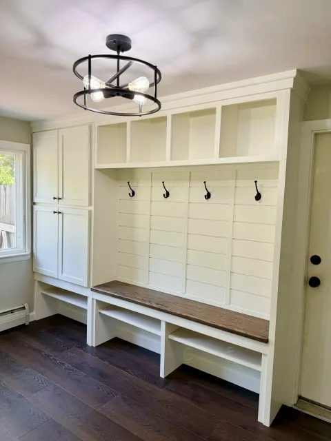 Kitchen mudroom with white built-in storage, black hooks, a wooden bench, a window, and a modern chandelier.