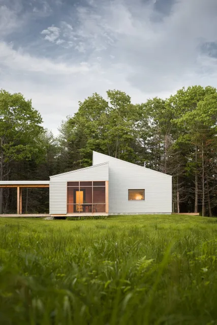 Modern house with white siding, featuring a screened porch with wooden framing, set on a grassy lawn with trees in the background and a partly cloudy sky overhead.