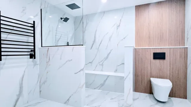 Modern bathroom remodel in Southington, CT with white marble walls, wood accent panel, black towel rack, and white toilet