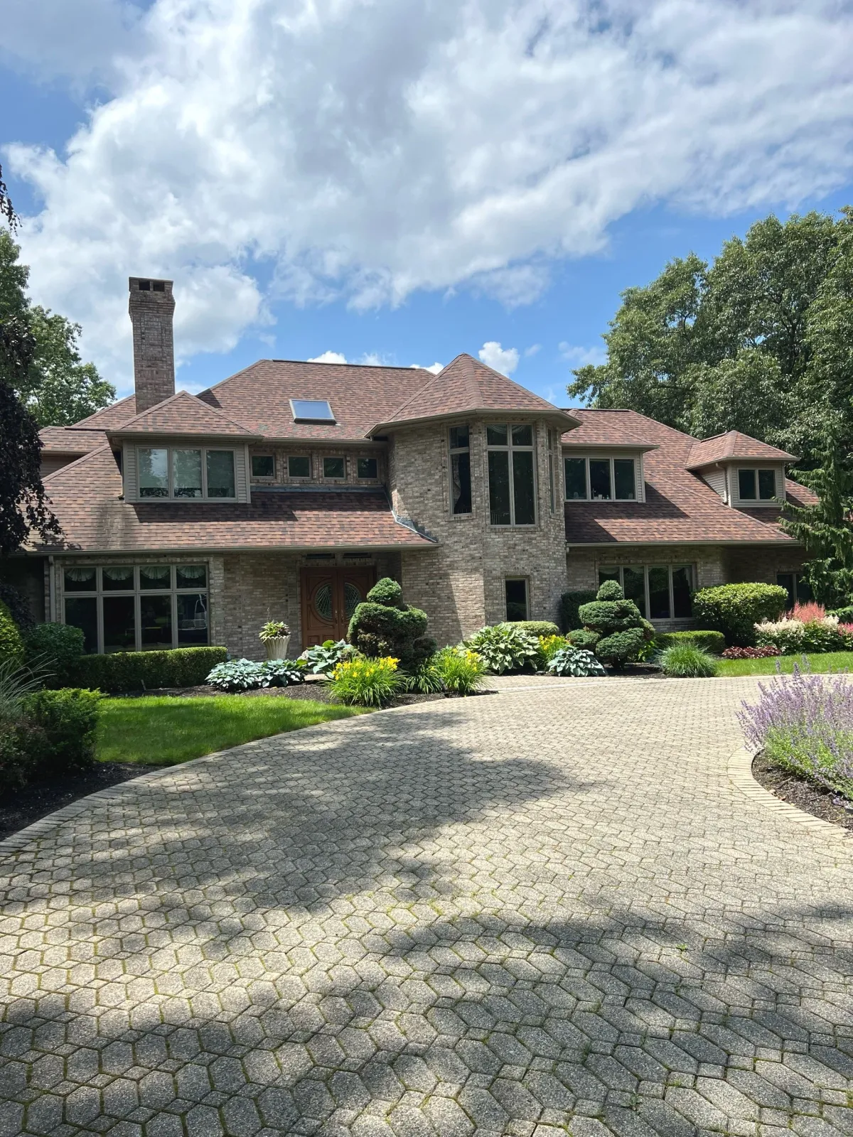 Large brick house with a brown roof, surrounded by a paved driveway and landscaped garden with various plants and shrubs, under a partly cloudy sky.