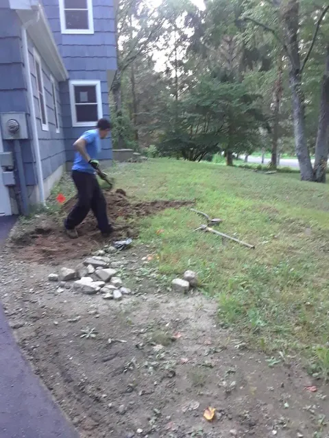 Person shoveling dirt and rocks beside a house in a grassy yard.