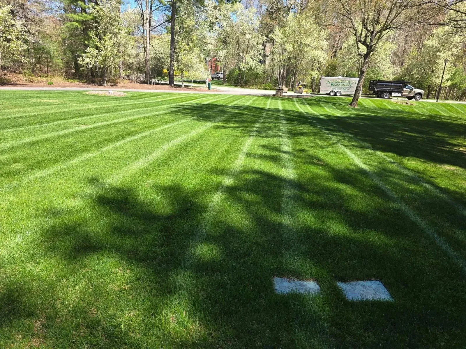 A lush, green lawn with distinct mowing stripes leading toward trees, a parked trailer, and a truck in the distance.