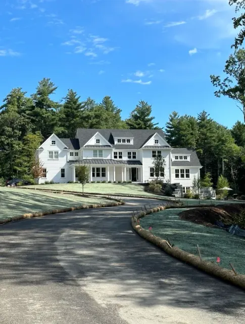 Large white mansion with a curved driveway, manicured lawn, and tall trees under a clear blue sky