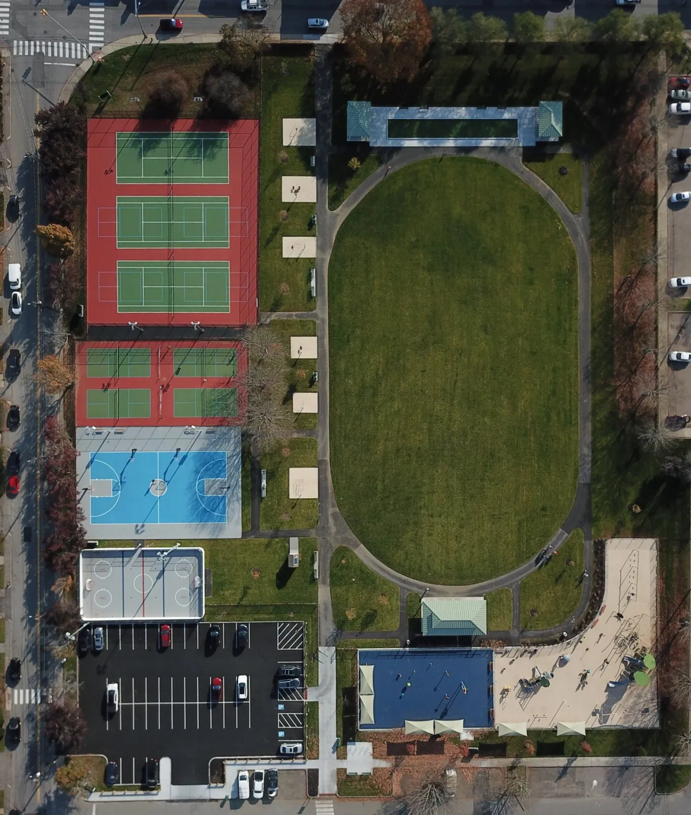 An aerial view of a community park featuring sports courts and an open grassy field. The park includes tennis courts, a basketball court, a skate park, and a playground, with parking lots and surrounding trees.