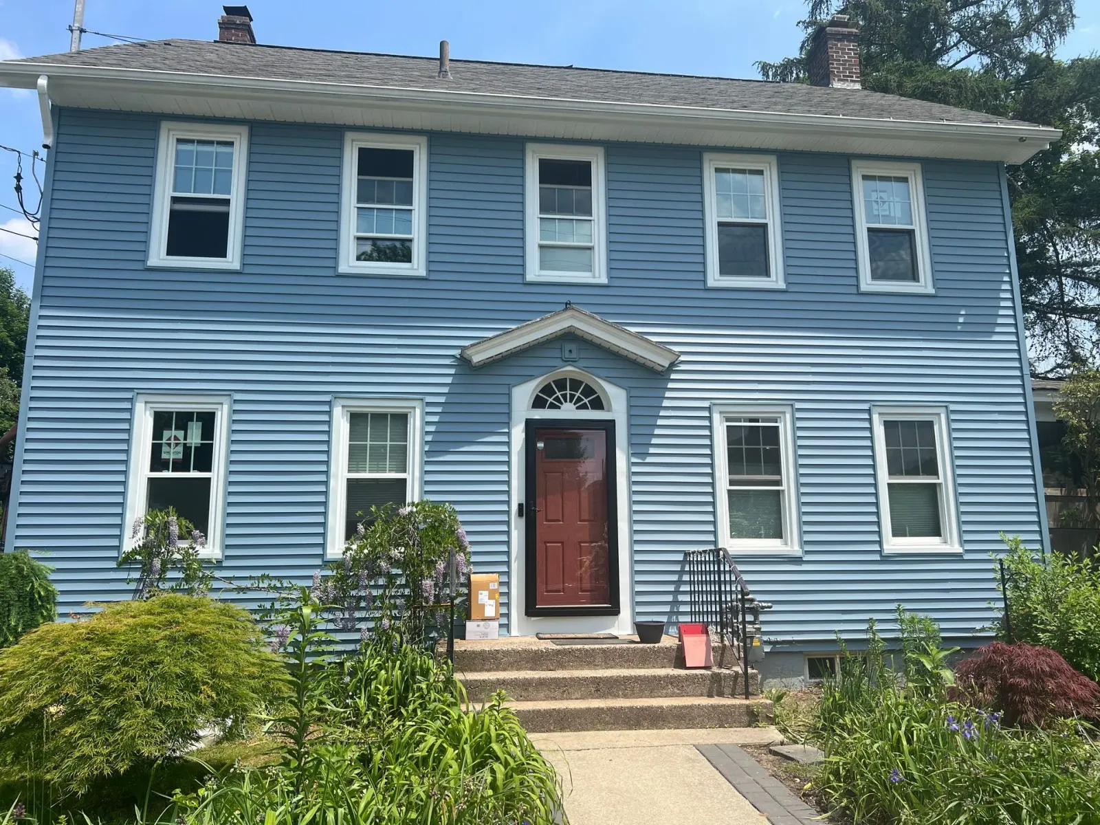 Blue two-story house with white trim, windows, and a red door, surrounded by greenery under a blue sky.