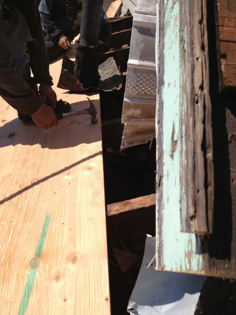 Worker kneeling on a wooden roof, handling boards beside a narrow gap and weathered siding.
