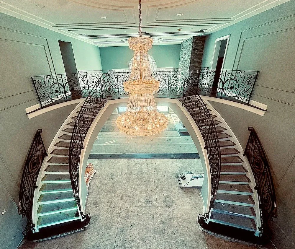 Elegant staircase with black wrought iron railings leading to a second floor, illuminated by a large crystal chandelier hanging above, in a spacious, nicely decorated interior.