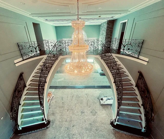 Elegant staircase with black wrought iron railings leading to a second floor, illuminated by a large crystal chandelier hanging above, in a spacious, nicely decorated interior.