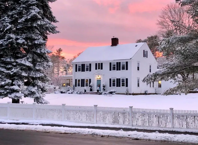 Snow-covered white house with black shutters, pink and orange sunset, white picket fence.