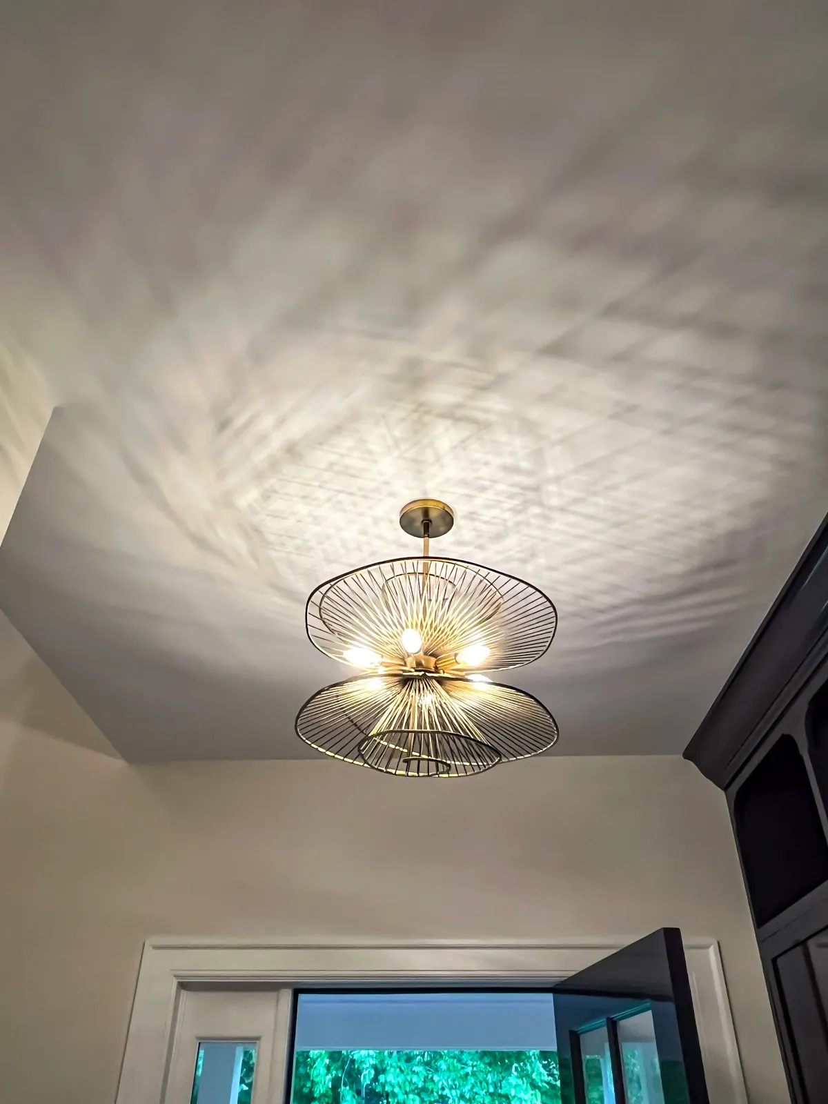 Modern ceiling light fixture with open wire design casting patterned shadows on the ceiling, above a doorway and window.