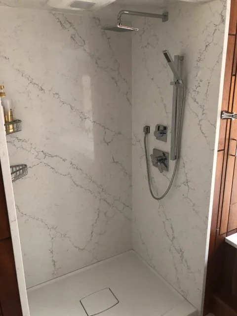 Modern shower with marble-patterned walls and chrome fixtures, including a rainfall showerhead and handheld shower wand. A small shelf holds toiletries.