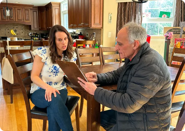 Two people sit at a wooden table in a kitchen. The man holds a clipboard and speaks to the woman.